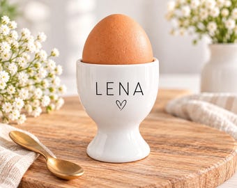 Op de afbeelding: Een witte keramieken eierdopje met de naam LENA in zwarte hoofdletters boven een klein zwart hartje. Een bruin ei staat in het eierdopje, dat een gladde, ronde voet en een uitlopende rand heeft om het ei stevig vast te houden.