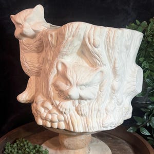 May include: A white ceramic planter shaped like a tree stump with two sculpted raccoons. One raccoon is perched on the rim, while the other is depicted peeking out from the center of the textured bark. The piece features a matte finish.