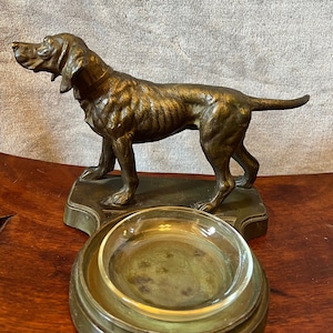 May include: A bronze sculpture of a dog standing on a base with a glass ashtray. The dog is facing left and has a collar around its neck.