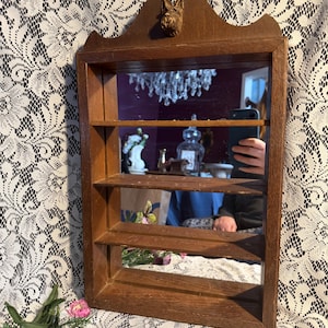 May include: A vintage wooden wall shelf with a mirror and three shelves. The frame is a warm brown color, and a small dog sculpture adorns the top. The mirror reflects a chandelier and other objects. The shelf is likely used for displaying small items.