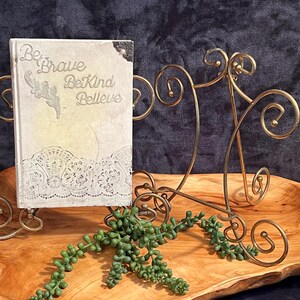May include: A decorative book with the words "Be Brave, Be Kind, Believe" on the cover, displayed on a gold metal stand. The book has a white lace design. The display is on a wooden tray with a green plant.
