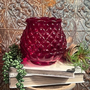 Może przedstawiać: Gliniany dzbanek w kolorze głębokiej czerwieni z fakturowanym, falistym wzorem. Dzbanek ma przezroczysty szklany uchwyt i stoi na stosie książek.