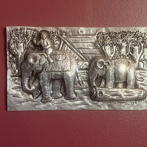 May include: A silver wall plaque depicting two elephants, one carrying a small figure on its back. The elephants are surrounded by trees and a small hut. The plaque is a traditional Thai design.