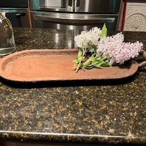 Puede incluir: Una bandeja marrón ovalada con asas, con flores de lilas. La bandeja está hecha de un material texturizado y se encuentra sobre una encimera moteada. Una botella de vidrio transparente está en el fondo.