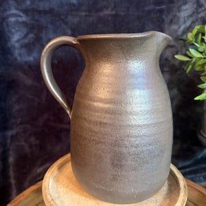 May include: A dark brown ceramic pitcher with a textured, matte finish and a rounded body. The pitcher features a curved handle on the left side and a small pouring spout at the top rim, showcasing a consistent earthy tone throughout the vessel.