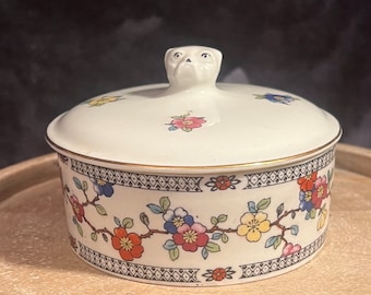 May include: A white porcelain trinket box with a floral design and a small dog figurine on the lid. The box has a gold trim around the top and bottom.