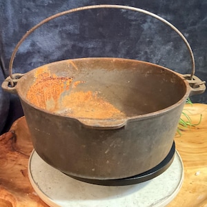 May include: A well-worn, cast iron Dutch oven with a curved metal handle. The pot shows signs of rust and age, with a dark, weathered exterior. The handle arches over the top, and the pot sits on a round base.