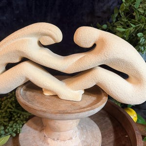 Op de afbeelding: Een beige, getextureerd beeldhouwwerk dat twee abstracte menselijke figuren in een zittende, verstrengelde positie weergeeft. De figuren zijn verbonden bij de armen en benen, wat een vloeiende vorm creëert met een ruwe afwerking.