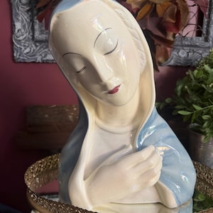 May include: A ceramic bust of a figure with closed eyes, featuring a cream-colored face and a light blue head covering that drapes over the shoulders. The figure has hands crossed over the chest, a smooth glossy finish, and subtle facial features.