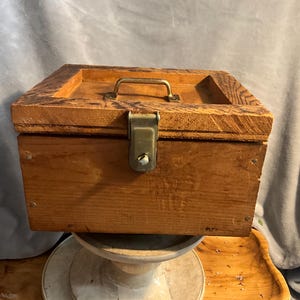 Könnte beinhalten: Eine Vintage-Holzkiste mit Messinggriff und Verschluss. Die Kiste ist rechteckig mit einem leicht erhöhten Deckel und besteht aus hellbraunem Holz. Der Griff ist gebogen und der Verschluss ist mittig angebracht.