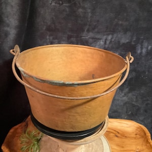 Może przedstawiać: Zabytkowe miedziane wiadro z metalowym uchwytem. Wiadro ma rustykalny wygląd z lekko przyciemnionym wykończeniem. Uchwyt jest przymocowany metalowymi pętlami. Wiadro stoi na drewnianym stojaku.