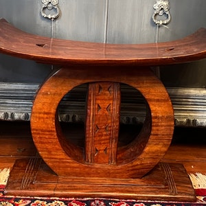 May include: A handcrafted wooden stool with a curved top and a circular design in the center. The stool is made of dark brown wood and features carved details. The base is rectangular with carved lines. The stool is a unique piece of furniture.