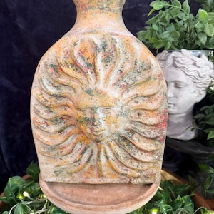 May include: A flat, rounded ceramic vase featuring a raised relief of a sun face with radiating rays. The surface has a weathered, mottled finish in shades of muted orange, yellow, green, and brown, with a narrow neck and a wide, circular base.