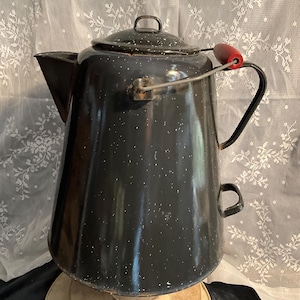 Puede incluir: Una cafetera vintage de esmalte negro con motas blancas. Tiene tapa, boquilla y asa con empuñadura de madera roja. La cafetera está sobre una bobina de madera.