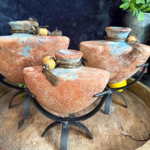 Op de afbeelding: Drie terracotta vaten met een verweerde, roodbruine en blauwachtige afwerking. Elk vat heeft een smalle hals omwikkeld met donker touw en een gele kraal, en rust op een zwarte metalen standaard met vier gebogen poten.