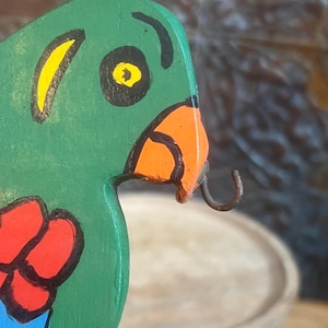 May include: Close-up of a wooden parrot figurine with green, red, blue, yellow, and orange paint. The parrot has a black eye and a hook on its beak.