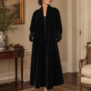 May include: A floor-length black velvet duster coat featuring a shawl collar, long sleeves with textured cuffs, and side pockets. The garment is worn over a simple black top and is paired with black pointed-toe heels.