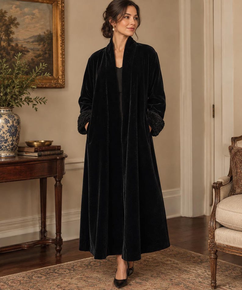 May include: A floor-length black velvet duster coat featuring a shawl collar, long sleeves with textured cuffs, and side pockets. The garment is worn over a simple black top and is paired with black pointed-toe heels.