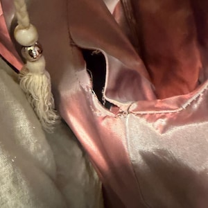 May include: A dusty rose satin garment featuring a visible seam opening along the side. A braided cord drawstring with a white wooden bead and a metallic silver bead hangs from the upper portion of the fabric, which has a smooth, reflective finish.
