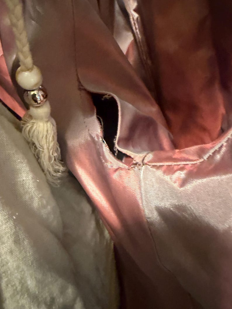 May include: A dusty rose satin garment featuring a visible seam opening along the side. A braided cord drawstring with a white wooden bead and a metallic silver bead hangs from the upper portion of the fabric, which has a smooth, reflective finish.