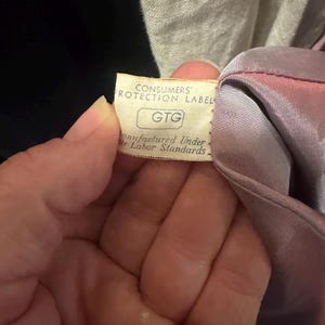 May include: A cream-colored fabric tag attached to a mauve garment. The tag features the text CONSUMERS' PROTECTION LABEL, the letters GTG inside a rectangle, and the phrase Manufactured Under Fair Labor Standards printed in black ink.