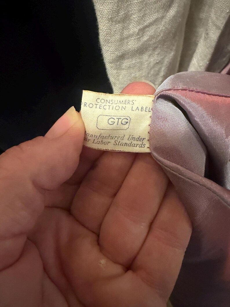 May include: A cream-colored fabric tag attached to a mauve garment. The tag features the text CONSUMERS' PROTECTION LABEL, the letters GTG inside a rectangle, and the phrase Manufactured Under Fair Labor Standards printed in black ink.