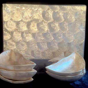 May include: Four white mother of pearl bowls stacked in two sets of two. The bowls are shaped like a fish with a pointed tail and a wide, rounded front. The bowls are sitting on a table in front of a larger white mother of pearl tray.