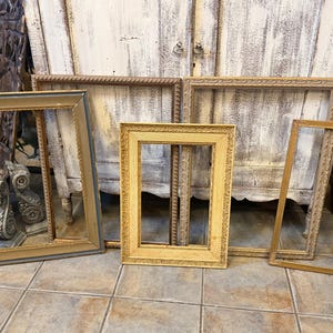 May include: A collection of four empty rectangular picture frames in various shades of gold and bronze. The frames feature ornate, textured, and carved detailing along their borders, showcasing different widths and decorative patterns.