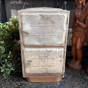 May include: Vintage metal two-door cooler with the word "CONSERVO" embossed on the top door. The cooler has a rectangular shape with a slightly rusted appearance. It features a handle on each door and sits on a metal base. The cooler is likely used for food storage.