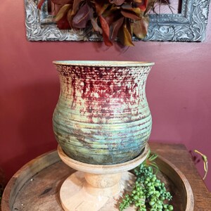 以下が含まれることがあります： 広がった縁と水平の溝がある陶器製の花瓶で、ティール、グリーン、ダークレッドの色合いが混ざり合ったアンティーク調の仕上げが施されています。花瓶は円形の土台を持つ明るい色の木製ペデスタルスタンドの上に置かれています。