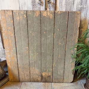 May include: A square wooden panel composed of six vertical planks with a distressed, weathered finish. The surface features layers of peeling sage green paint revealing natural wood tones underneath, creating a textured, aged appearance.