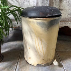 May include: A cylindrical metal trash can with a distressed cream-colored body featuring vertical drip marks and rust spots. It has a dark, weathered lid and a small white foot pedal at the base for opening the lid, showing signs of heavy wear.