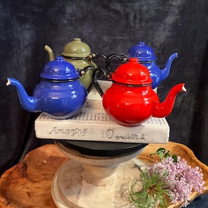 May include: Four colorful teapots in blue, green, and red sit atop a decorative stand. The teapots have black handles and spouts. The red teapot is in the foreground, with the text "Amazing Dreams" visible on a book beneath them.