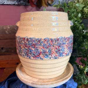 Puede incluir: Jardinera de cerámica de color crema con una superficie texturizada y una banda de diseño moteado azul y rojo. La jardinera tiene dos asas pequeñas y se asienta sobre un platillo a juego. Un artículo decorativo para plantas.