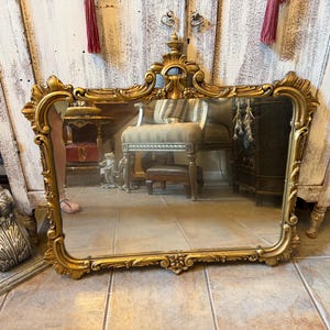 May include: A rectangular wall mirror featuring an ornate gold-toned frame with intricate scrollwork, floral carvings, and a decorative crest at the top center. The frame has a polished finish with detailed relief patterns along all four edges.