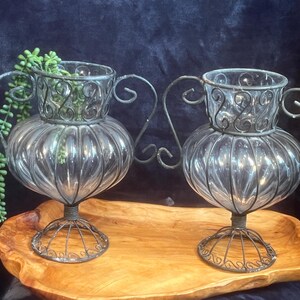 May include: Two clear glass vases with a decorative metal cage design and handles. The vases are sitting on a wooden tray.