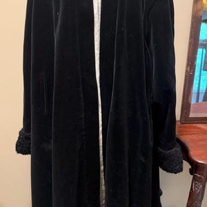 May include: A long black velvet coat featuring a shawl collar, long sleeves with textured black cuffs, and an open front design. The interior is lined with a dark maroon fabric, and the coat is displayed on a mannequin over a white garment.