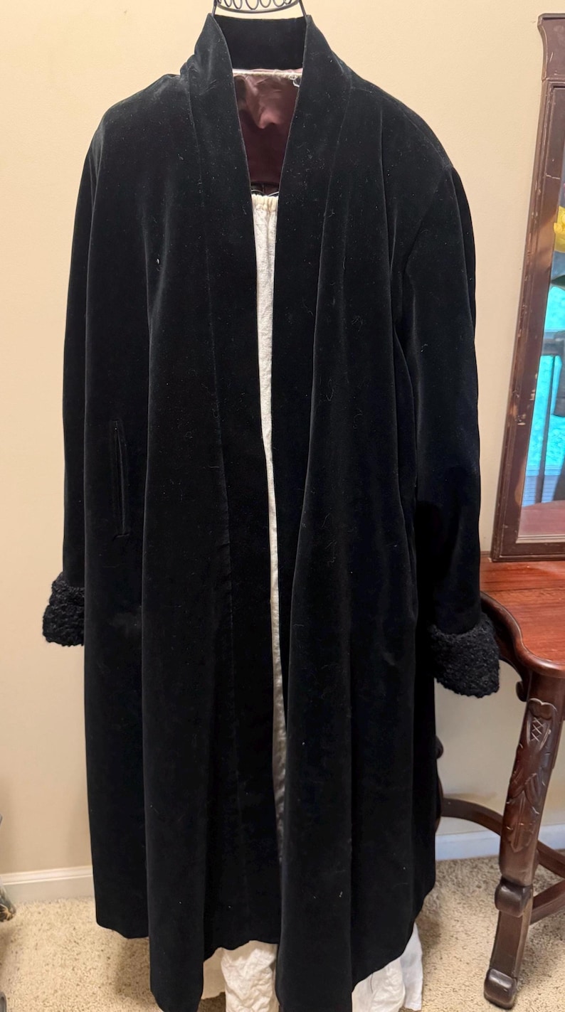 May include: A long black velvet coat featuring a shawl collar, long sleeves with textured black cuffs, and an open front design. The interior is lined with a dark maroon fabric, and the coat is displayed on a mannequin over a white garment.