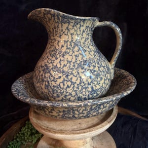 May include: A ceramic jug and matching basin set featuring a cream-coloured base with a blue sponge-painted or speckled pattern. The jug has a curved handle and a pouring spout, while the basin has a wide, shallow, circular shape.