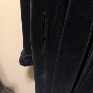 May include: A long black velvet coat featuring a vertical slit pocket on the side and a textured, curly black faux fur cuff on the sleeve. The garment has a smooth, matte black finish and a structured, full-length silhouette.