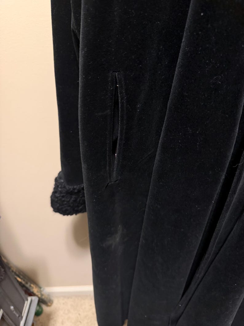 May include: A long black velvet coat featuring a vertical slit pocket on the side and a textured, curly black faux fur cuff on the sleeve. The garment has a smooth, matte black finish and a structured, full-length silhouette.
