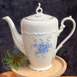 Puede incluir: Tetera de porcelana blanca con un diseño floral con flores azules de nomeolvides. La tetera tiene una tapa con un pomo decorativo y un borde dorado alrededor del borde.