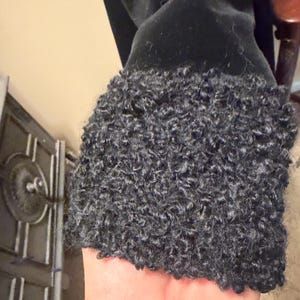 May include: A black garment sleeve featuring a thick, textured cuff made of dark gray curly faux fur material. The main body of the sleeve is composed of a smooth, solid black fabric that contrasts with the dense, looped pile of the cuff.