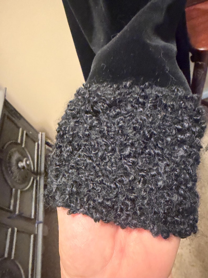 May include: A black garment sleeve featuring a thick, textured cuff made of dark gray curly faux fur material. The main body of the sleeve is composed of a smooth, solid black fabric that contrasts with the dense, looped pile of the cuff.