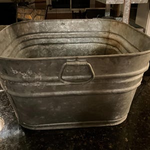 May include: A rectangular galvanized metal tub with a slightly weathered appearance. The tub has a series of horizontal ridges and a handle on one side. The overall color is a muted gray, suggesting an aged or vintage aesthetic.