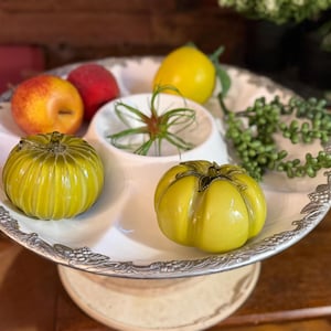 Op de afbeelding: Een wit en zilver serveerplateau met een decoratieve rand, met diverse kunstmatige groenten en fruit. De opstelling omvat gele tomaten, een citroen, een appel en een rode vrucht, samen met wat groen.