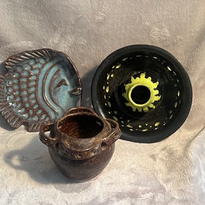 May include: Three ceramic pottery pieces: a blue and brown fish-shaped dish, a brown pot with two handles, and a black bowl with a yellow center and white dots.
