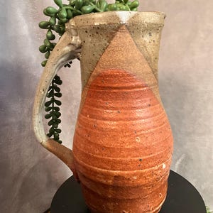 May include: Handmade ceramic pitcher with a textured, reddish-brown body and a tan upper section. The pitcher features a large handle and is adorned with a green plant. The design is rustic and unique, suitable for home decor.