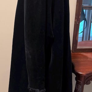 May include: A long, black velvet coat featuring a high collar and long sleeves with textured, dark, fuzzy cuffs. The coat has a smooth, matte finish and hangs in a straight, structured silhouette, extending down to the lower portion of the frame.