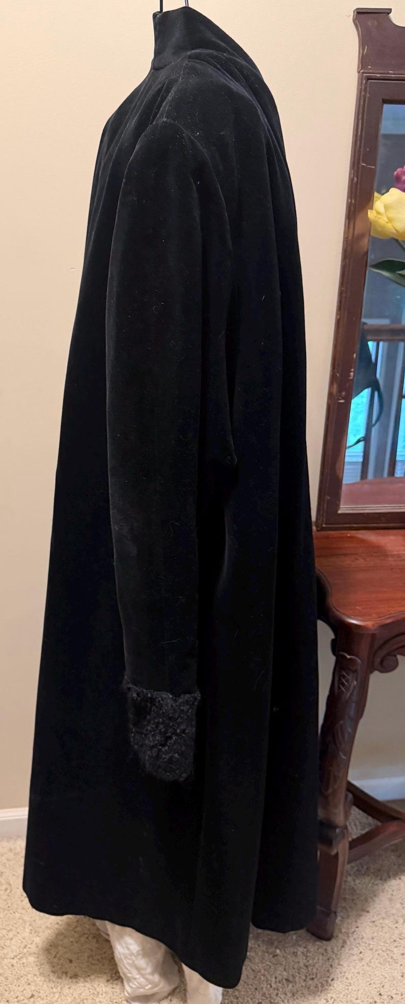 May include: A long, black velvet coat featuring a high collar and long sleeves with textured, dark, fuzzy cuffs. The coat has a smooth, matte finish and hangs in a straight, structured silhouette, extending down to the lower portion of the frame.